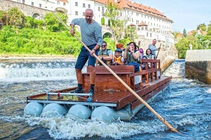 A traditional wooden raft glides gently down the Vltava, steered by a raftsman with a long pole through the calm current.